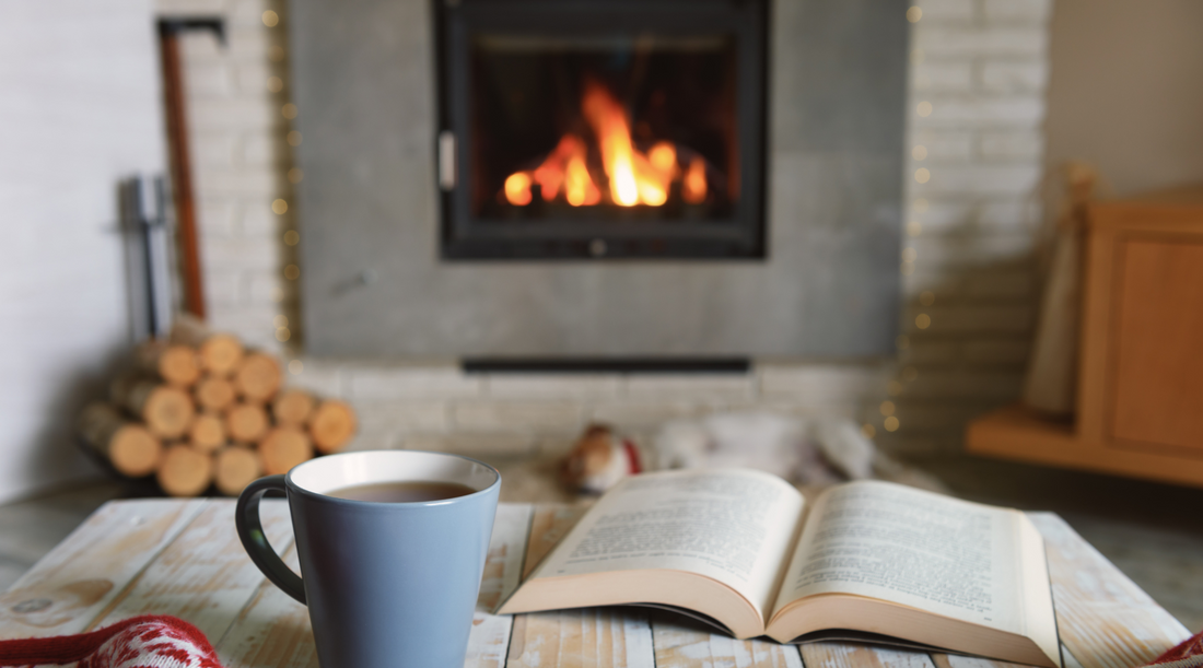 cozy setting in room with fireplace in the background and mug of tea with open book placed on table in foreground