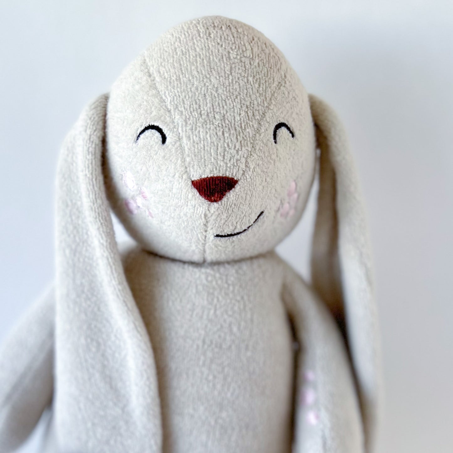 close up on face of a greige plush bunny toy with embroidered features, brown nose, and pink patches of eczema