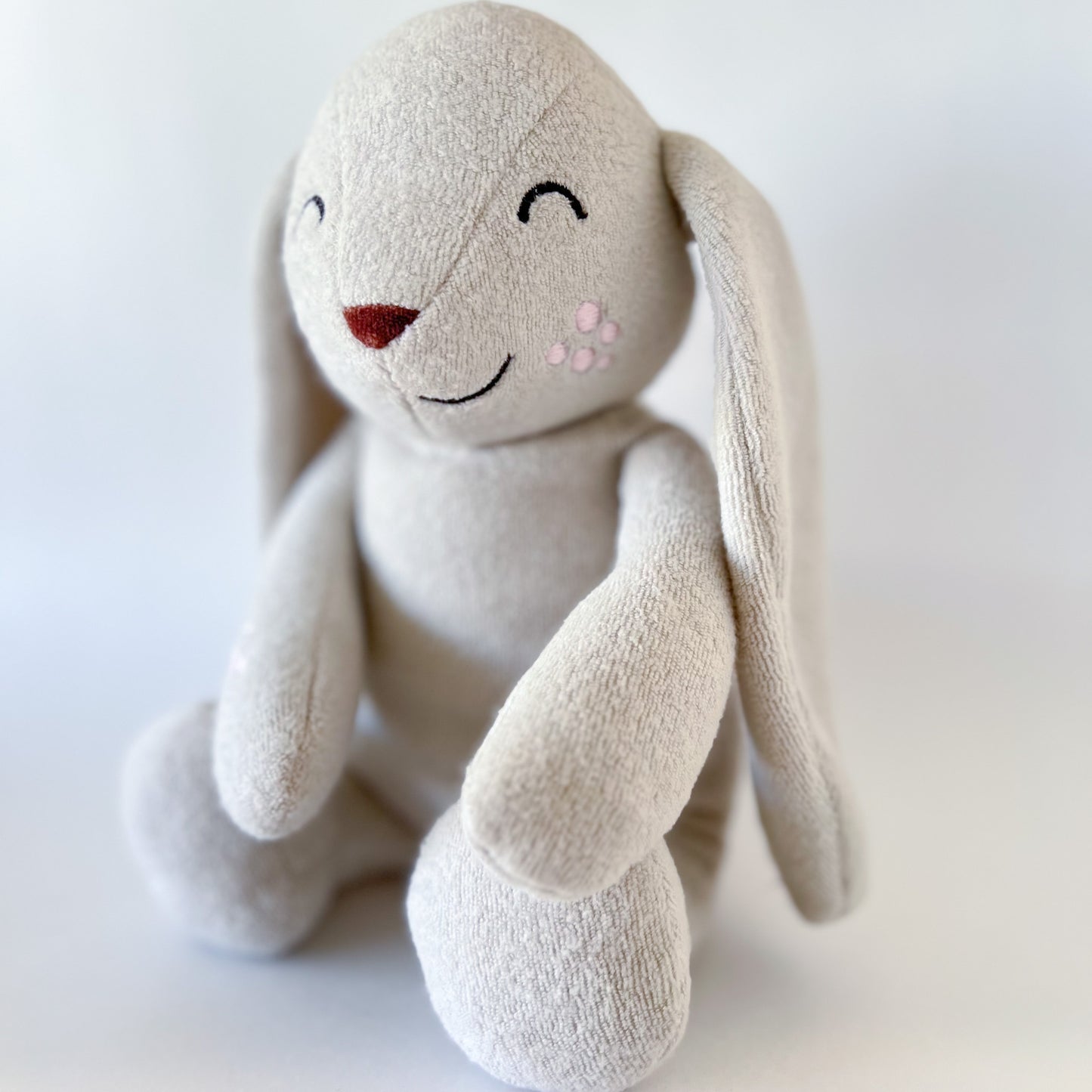 side angle shot of a light grey stuffed animal bunny sittting up with long ears, brown nose, black emroidered facial features, and pink spots on cheek