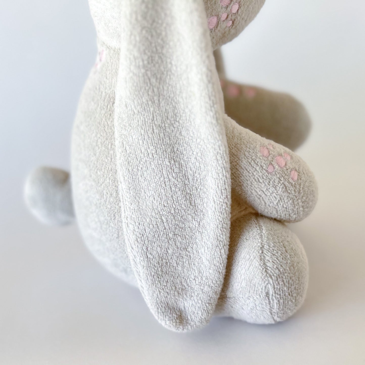 side view of off white plush toy bunny showing long ears, bunny tail, and pink embroidered patches on cheek and arms